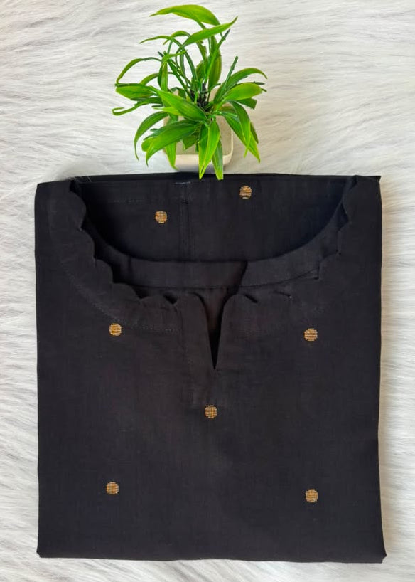 A folded black kurta with a scalloped neckline and small gold button accents, displayed on a white faux fur surface with a small green plant above it.